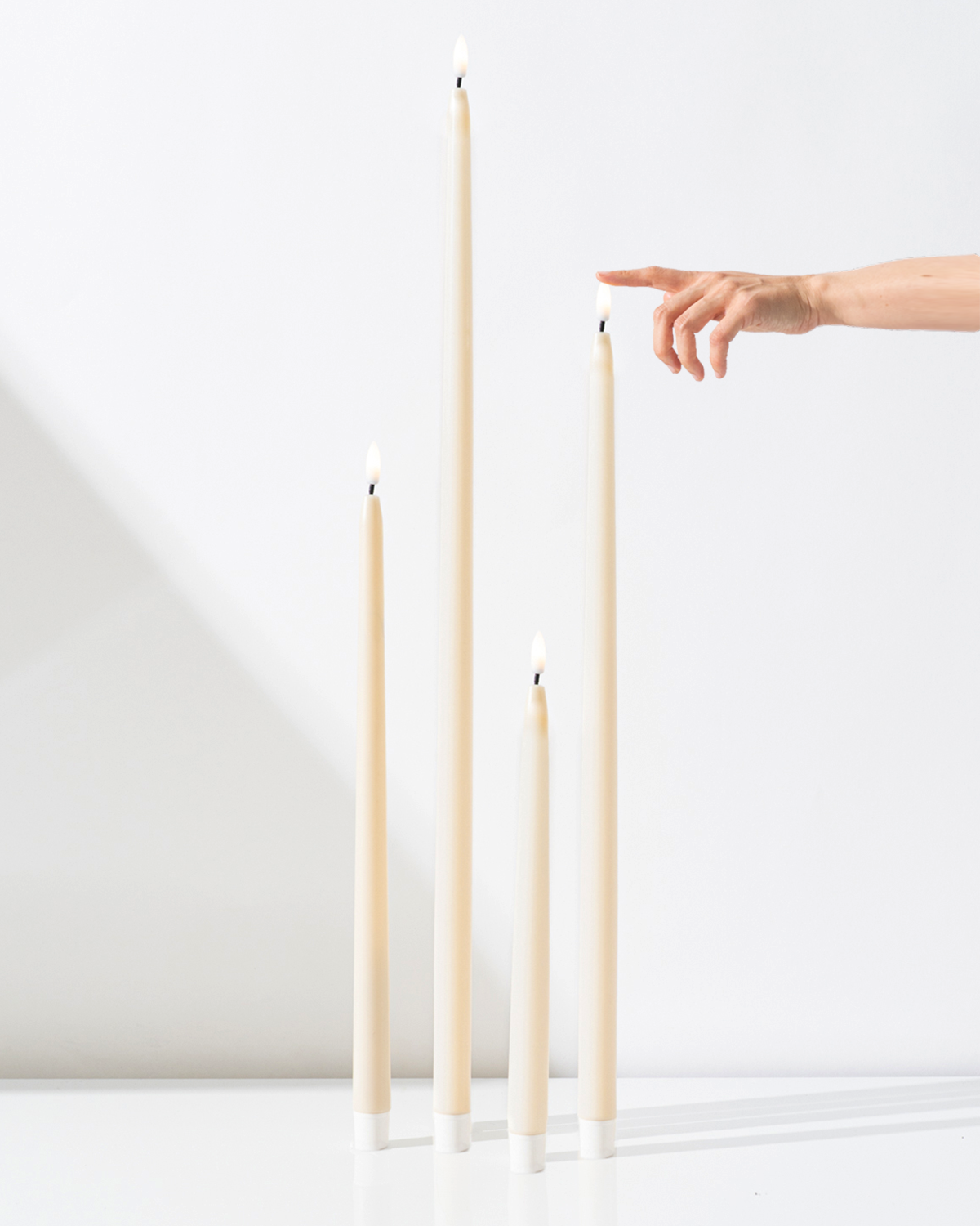 Four Hall of Flame Ivory Ultra-Bright LED Flameless Taper Candles of various heights stand upright on a white surface, while a hand with an extended finger reaches toward the tallest one. The background is plain white.