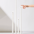 Four Hall of Flame White | Realistic LED Flameless Taper Candles of different heights stand on a white surface. A hand enters from the right to touch the tallest candle’s lifelike flame.