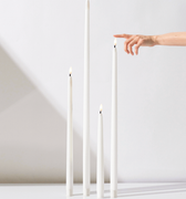 Four Hall of Flame White | Realistic LED Flameless Taper Candles of different heights stand on a white surface. A hand enters from the right to touch the tallest candle’s lifelike flame.
