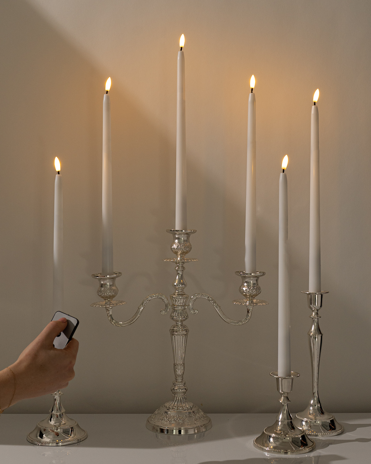 A hand holds a Hall of Flame Remote for Ultra-Bright LED Taper Candles near a silver candelabra with two lit white LED candles, set against a plain light-colored background.