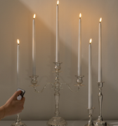 A hand holds a Hall of Flame Remote for Ultra-Bright LED Taper Candles near a silver candelabra with two lit white LED candles, set against a plain light-colored background.