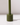 The Hall of Flame Moss Green Wax Candle Holder, a sustainable plant-based cylindrical piece with a tall, thin stem, stands on a white surface casting a faint rightward shadow against a softly lit plain background.