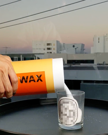 Someone pours The Bug-off! series | SANDWAX Scented Candle Refill by Hall of Flame from a large yellow container into a glass with a pixelated label, on a black tray near a window overlooking buildings at sunset.