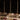 A formal dinner table features a white tablecloth, three Hall of Flame ULTRABRIGHT™ LED Flameless Taper Candles in gold holders, glassware, cutlery, and hardcover books with menus at each place. The softly lit background is dark.