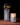 Two Hall of Flame Spike Hand Blown Vases sit on a brown surface against a dark backdrop—one holds dark purple and green flowers, the other glows warmly with a lit candle inside.