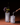 A hand pours small white pebbles into the Hall of Flame Spike Hand Blown Vase next to another vase with purple and yellow flowers. Both rest on a brown surface against a dark background.