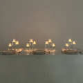 The Hall of Flame Floating Tealight Wedding Bundle adds a warm, soft glow to a minimal dining table set for three, with glass vessels and lit candles—an elegant wedding centrepiece for any plain background.