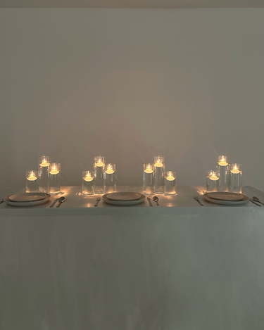 The Hall of Flame Floating Tealight Wedding Bundle adds a warm, soft glow to a minimal dining table set for three, with glass vessels and lit candles—an elegant wedding centrepiece for any plain background.