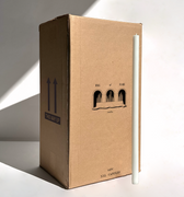 A tall white candle stands next to a large Hall of Flame box labeled “Bulk Candles | Wholesale by-the-box – 60 PC XXL” with “THIS WAY UP” arrows, ideal for wholesale candles Australia. Light from the left casts shadows.