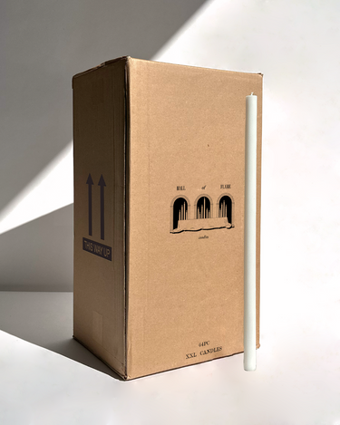 A tall white candle stands next to a large Hall of Flame box labeled “Bulk Candles | Wholesale by-the-box – 60 PC XXL” with “THIS WAY UP” arrows, ideal for wholesale candles Australia. Light from the left casts shadows.