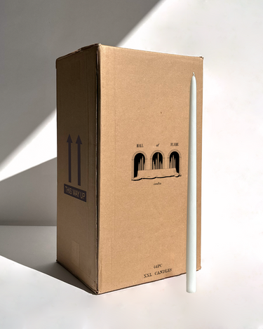 A tall white candle stands upright beside a large Hall of Flame box labeled Bulk Candles | Wholesale by-the-box, featuring three arches and the words THIS WAY UP—ideal for anyone seeking bulk or wholesale candles in Australia.