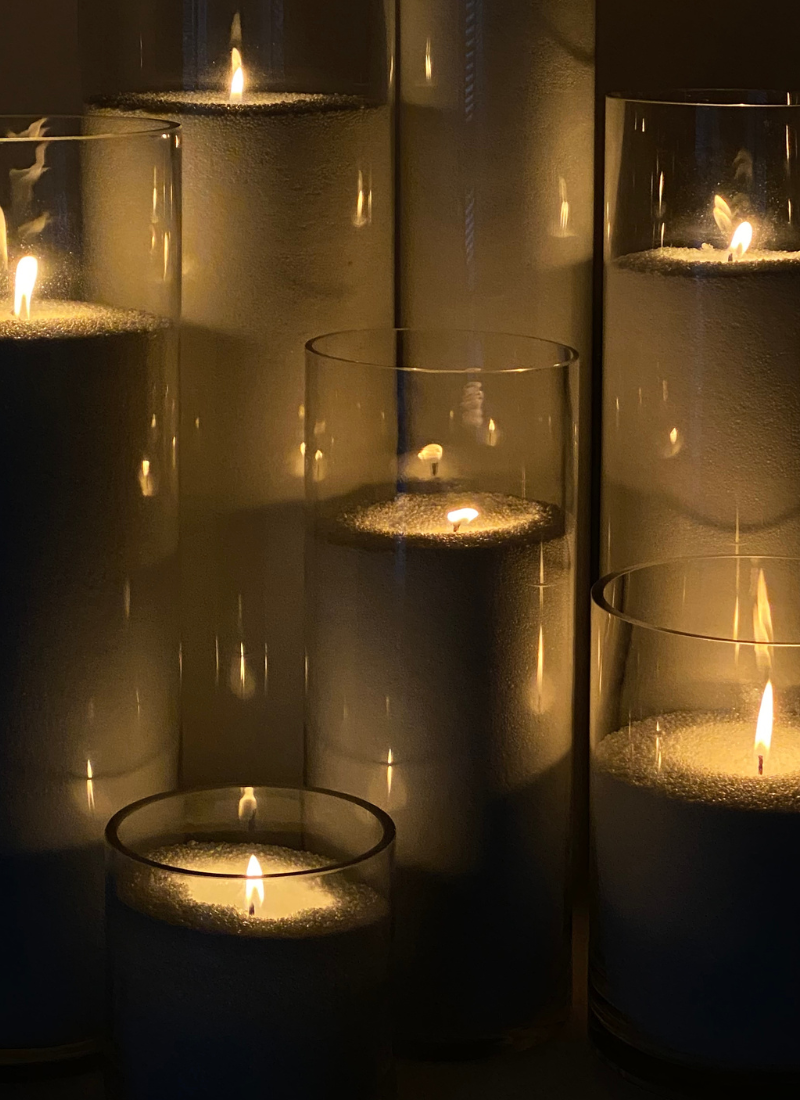 Several tall, clear glass vases filled with white sand each hold a candle burning with a soft, warm flame, creating a calm and cozy atmosphere in low light.