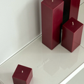 Four burgundy square candles of varying heights sit unlit on a glossy white Acrylic Candle Drip Tray by Hall of Flame near a wall, their reflections visible on the tray’s surface.