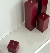 Four burgundy square candles of varying heights sit unlit on a glossy white Acrylic Candle Drip Tray by Hall of Flame near a wall, their reflections visible on the tray’s surface.