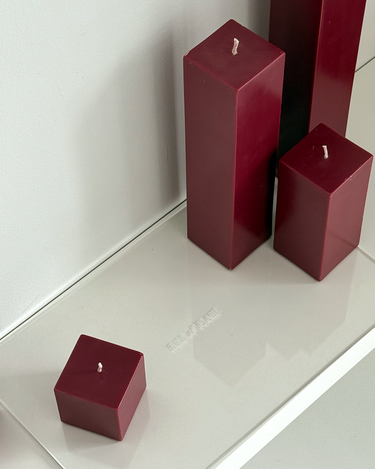Four burgundy square candles of varying heights sit unlit on a glossy white Acrylic Candle Drip Tray by Hall of Flame near a wall, their reflections visible on the tray’s surface.