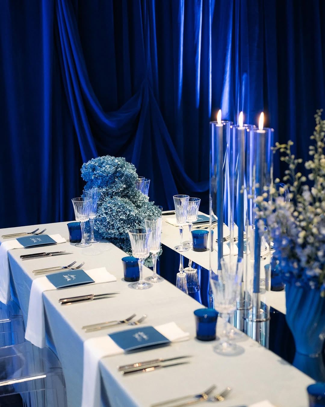 A formal dining table set with white tablecloths, blue napkins, crystal glasses, silver cutlery, blue flower arrangements, and tall glass candle holders with lit candles, against rich blue draped curtains.