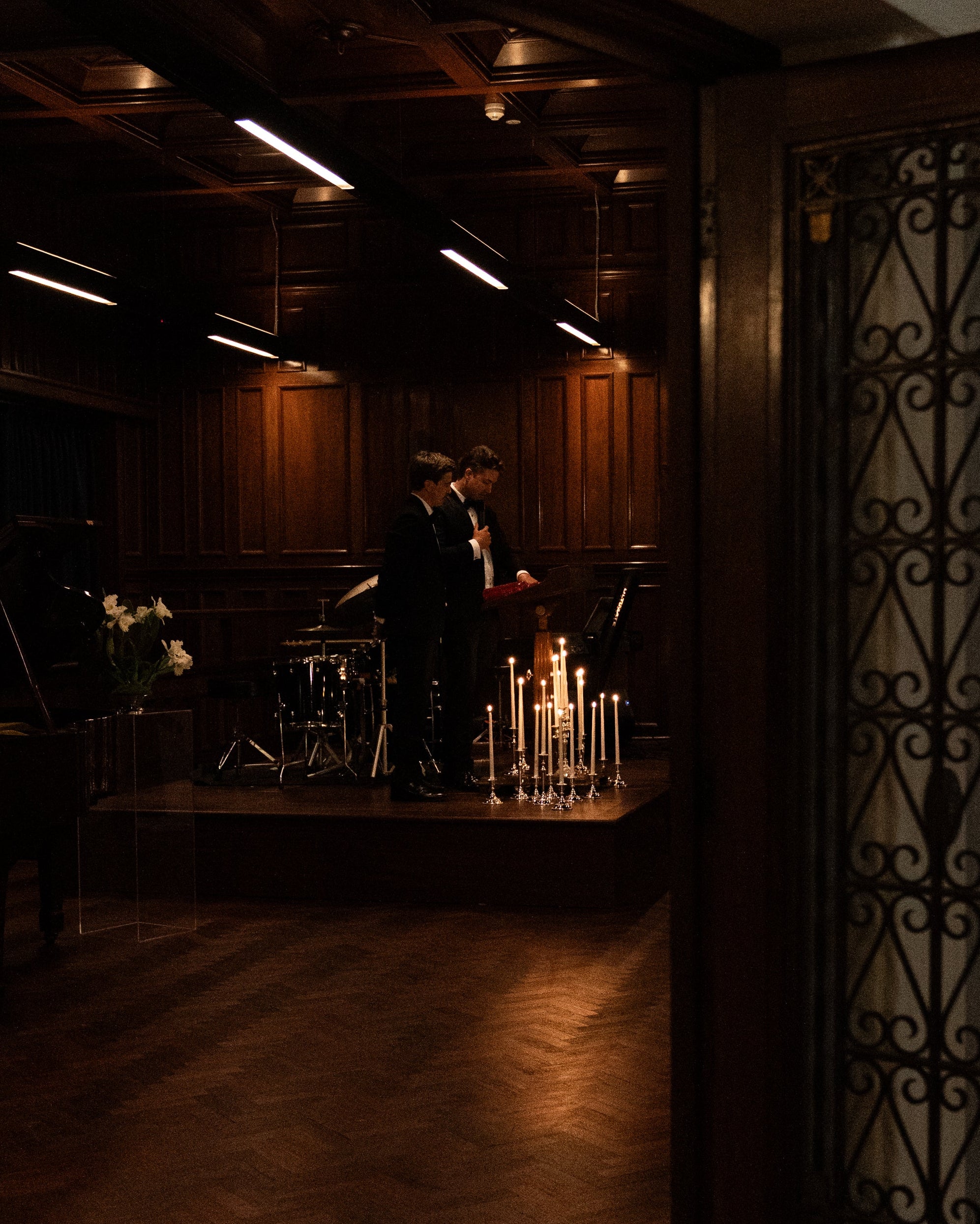 Two people in suits stand on a wooden stage with a drum set and piano nearby. Hall of Flame ULTRABRIGHT™ LED Flameless Taper Candles are arranged in front, creating a warm, intimate atmosphere without open flames.