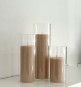 The Hall of Flame Pillars in Glass Vase Trio features three beige pillar candles of different heights arranged in clear glass holders on a white surface against a white wall.