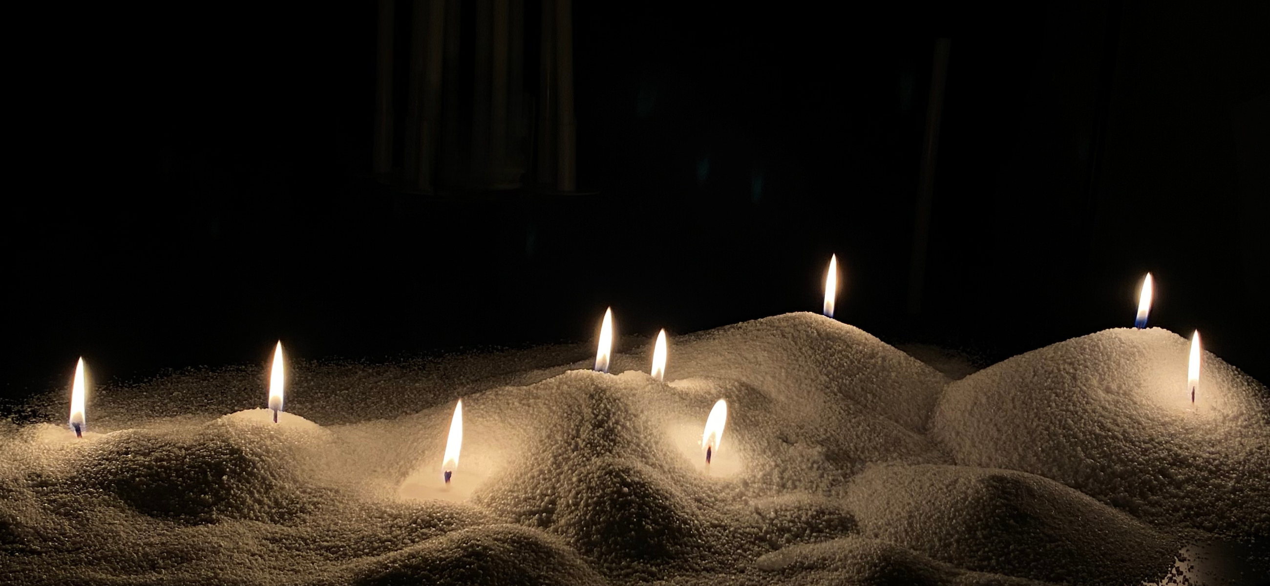Seven lit candles are placed upright in small mounds of white granules, possibly salt or sugar, against a dark background, creating a serene, softly illuminated scene.